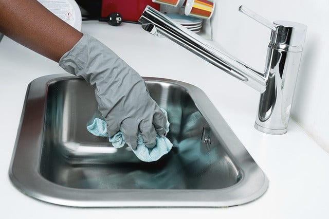 Hand im Gummihandschuh reinigt ein Spülbecken aus Chromstahl mit einem Tuch